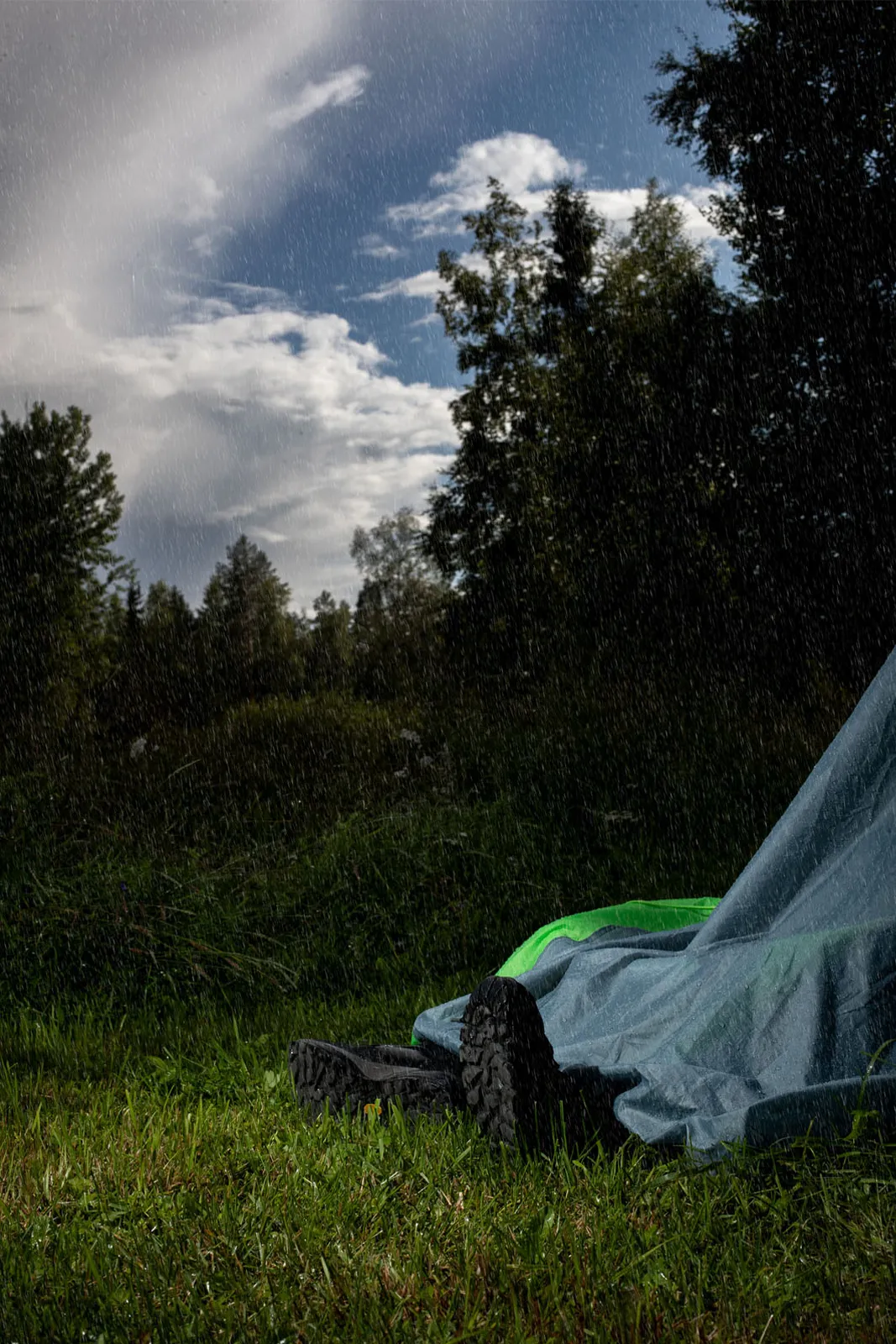 Mies makaa romahtaneessa teltassa sateessa. Vain hänen kenkänsä näkyvät teltan alta.